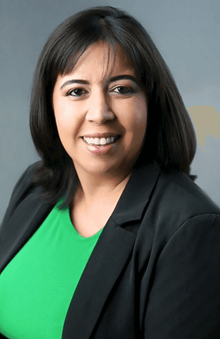 Person with shoulder-length dark hair wearing a green top and black blazer against a neutral background.